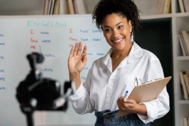 Professora sorridente em frente a um quadro com lições de inglês, acenando para uma câmera, representando aulas de idiomas presenciais e online.